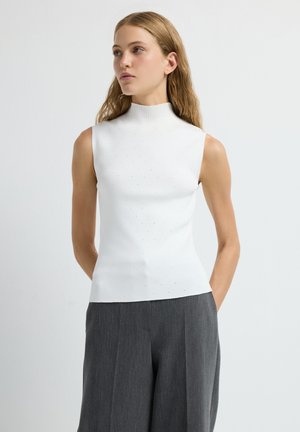 Jeune femme aux longs cheveux portant un haut blanc sans manches et un pantalon large gris, se tenant avec les mains derrière le dos.