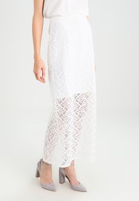 Gonna maxi bianca in pizzo con motivo floreale, tessuto semi-trasparente, foderata sotto, abbinata a scarpe con tacco alto grigie.