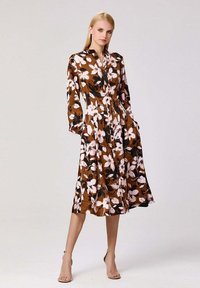 Robe midi florale marron avec manches longues, devant à boutons et jupe évasée. Présente de grandes fleurs roses et des feuilles vertes sur un fond marron.