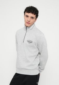 Γκρι φούτερ με φερμουάρ και ψηλό γιακά, με λογότυπο στο στήθος, υφή υφάσματος και άνετη εφαρμογή. Φοριέται με μαύρο παντελόνι κάτω.