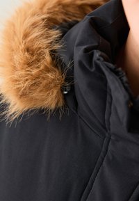 Czarna kurtka z kapturem wykończonym futrem, z beżowym futrzanym obszyciem. Zbliżenie pokazuje teksturę materiału oraz szczegóły guzików na kołnierzu.