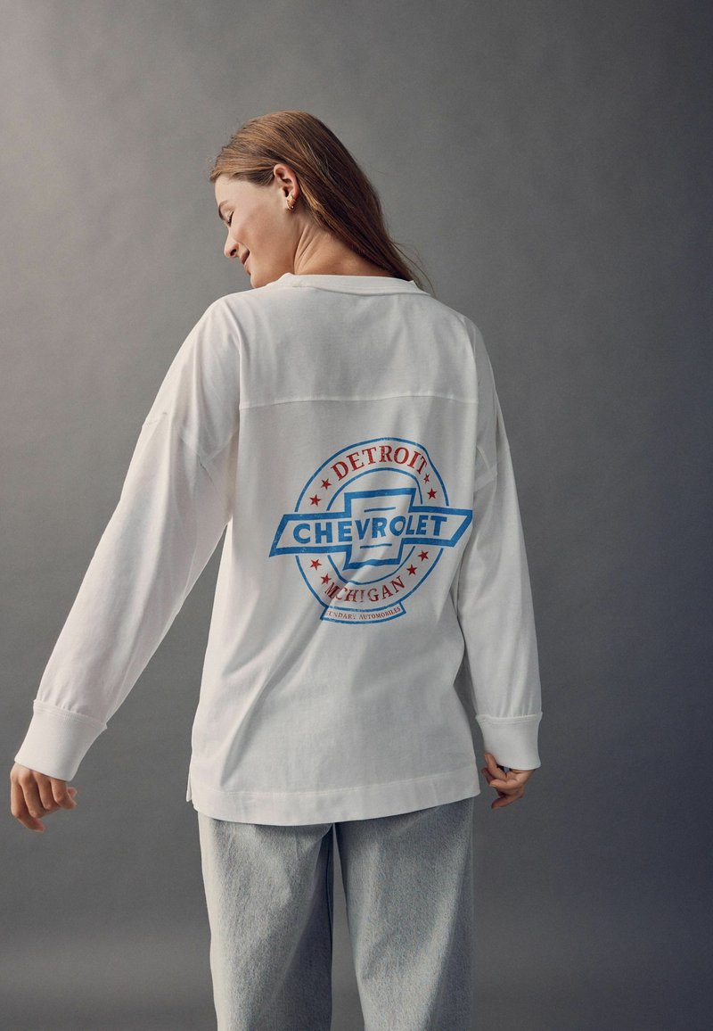 Tricou alb cu mânecă lungă, prezentând un logo Chevrolet albastru și roșu, cu textul "Detroit Michigan". Țesătura pare moale și are o croială lejeră.
