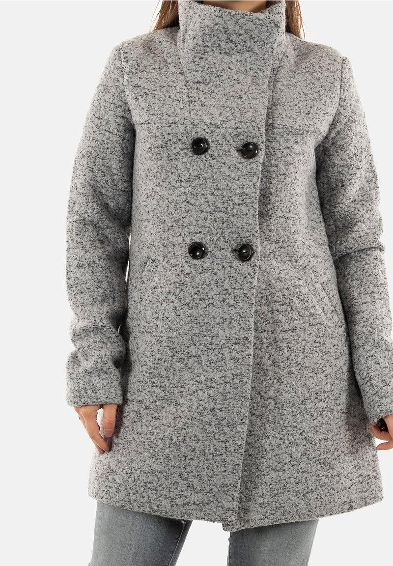 Femme portant un manteau gris texturé à double boutonnage avec quatre boutons noirs et des poches latérales, associé à un pantalon gris.