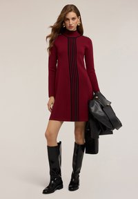 Šaty po kolena v barvě burgundy se černými vertikálními pruhy, s vysokým límcem a dlouhými rukávy. Modelka drží černou koženou bundu. Černé vysoké kozačky.
