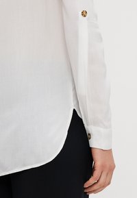 Witte lange mouwen shirt met een gladde textuur, openingen aan de zoom en een met knoop gesloten manchet met een ronde, contrasterende knoopdetail.
