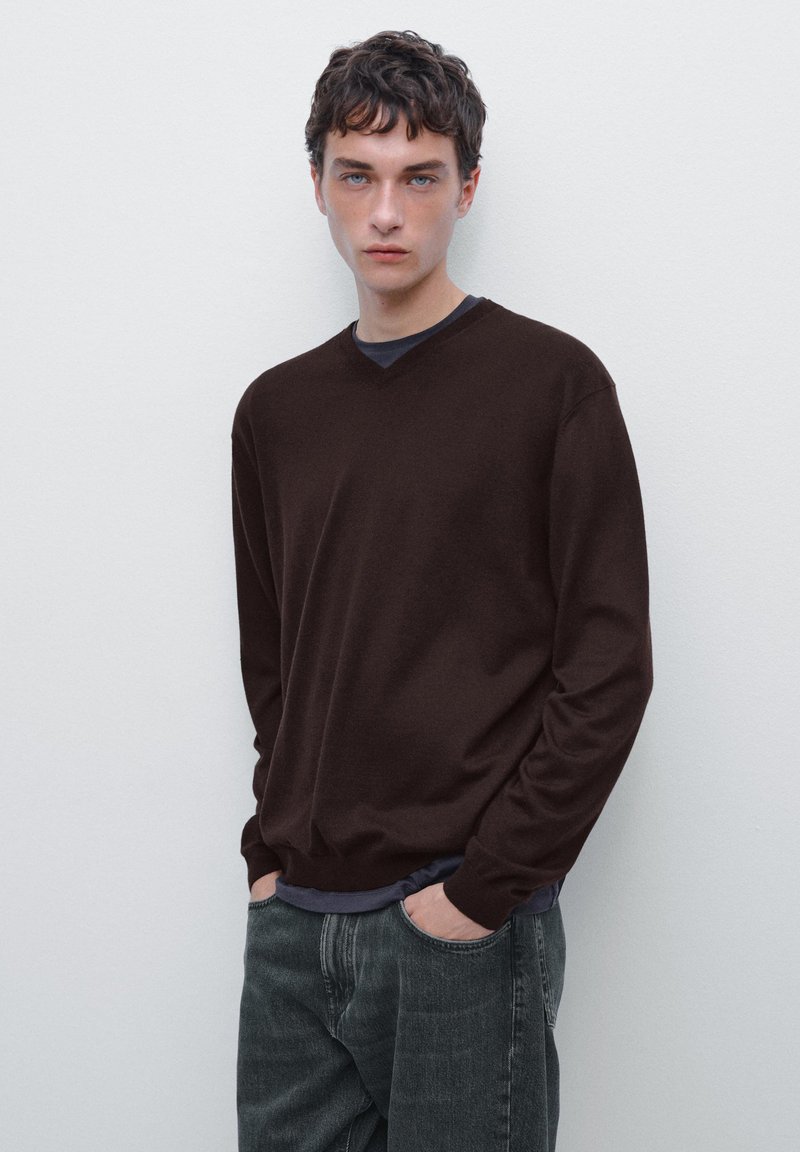 Brauner V-Ausschnitt-Pullover mit langen Ärmeln, aus einem glatten Material, über einem dunkelblauen T-Shirt getragen. Gestylt mit grauen Jeans.