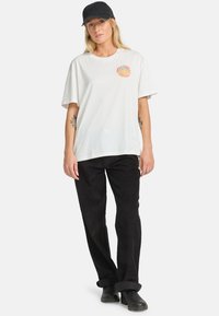 T-shirt bianco in cotone di taglio largo con grafica circolare arancione; abbinato a pantaloni in corduroy neri e cappellino a visiera piatta nero.