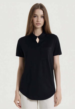 Giovane donna con capelli lunghi e lisci che indossa una camicetta nera a maniche corte con scollatura a goccia e pantaloni bianchi, in piedi davanti a uno sfondo semplice.