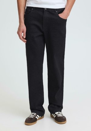 Mann trägt gerade schwarze Jeans und braune Sneakers mit weißen Streifen, steht mit einer Hand in der Tasche vor hellem Hintergrund.