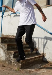 Czarne spodnie i biała koszulka z kieszonką na piersi, noszone z czarnymi butami, na schodach na świeżym powietrzu. Niebieska balustrada w tle.