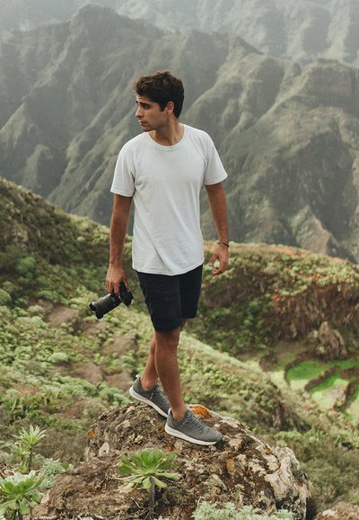 En person i en vit t-shirt och svarta shorts står på en sten och håller en kamera. De har på sig grå sneakers och omges av ett bergigt landskap.