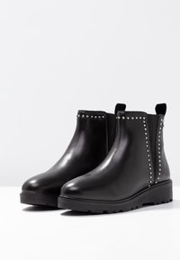 Botas negras de cuero hasta el tobillo con punteras redondas, paneles laterales elásticos y acentos de tachuelas plateadas a lo largo de las costuras. Suela de goma gruesa.