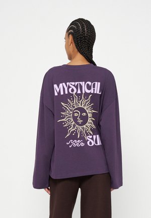 Personne aux cheveux tressés portant un t-shirt violet à manches longues avec un graphique de soleil et le texte "Mystical Sun" dans le dos.