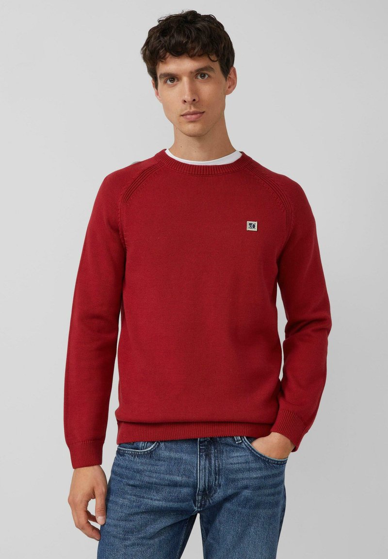 Junger Mann in einem roten Strickpullover mit kleinem Logo auf der Brust und blauen Jeans, der vor einem schlichten hellgrauen Hintergrund posiert.