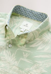Camicia verde chiaro a maniche corte con tessuto strutturato e stampe botaniche bianche. Design con collo e fodera interna blu con motivo.
