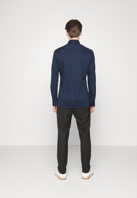 Långärmad, kroppsnära skjorta i mörkblått med ett texturerat geometriskt mönster. Parat med svarta byxor och vita sneakers.