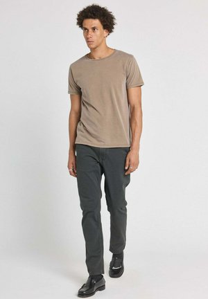 Homme aux cheveux bouclés marchant, portant un t-shirt beige à manches courtes, un pantalon gris foncé, des chaussettes noires et des mocassins noirs brillants.