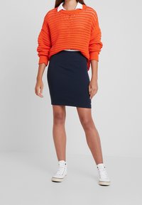 Oranžový pletený svetr s volným střihem, kombinovaný s dobře padnoucí námořnickou sukní a bílými vysokými teniskami. Hladká textura a jasné barvy.