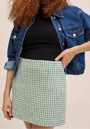 Femme portant une jupe tissée vert clair et blanche, un haut côtelé noir, et une veste en jean bleue aux manches retroussées.