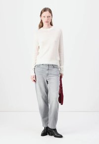 Kremfarget ribbestrikket genser, lysegrå jeans og svarte ankelstøvler. Modellen holder en burgunder veske. Nydelig, moderne og avslappet passform.