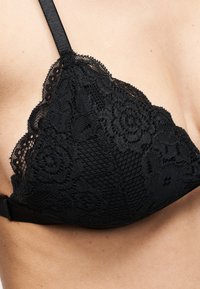 Μαύρο δαντελένιο σουτιέν με floral μοτίβα που φοριέται σε γυμνό δέρμα, με ρυθμιζόμενο λουράκι και κυματιστά άκρα.