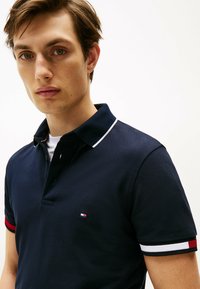 Polo navy in tessuto testurizzato, con colletto bianco a contrasto e strisce rosse e bianche sui maniche. Logo piccolo visibile.