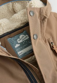 Waterproof jacket in tan, featuring a fluffy cream lining, blue accents, metal snaps, and a label with cloud graphics and text.