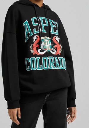 Person iført sort hættetrøje med turkis og hvid tekst "ASPEN COLORADO" og rødt ornamentalt design foran, kombineret med sorte bukser.