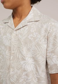 Košilové tričko s krátkým rukávem v béžové tkanině s bílým listovým květinovým vzorem, s klopovým límcem a lehkým zdobením knoflíků.