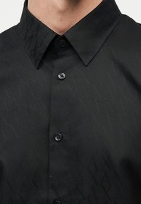 Κοντινό πλάνο ενός ατόμου που φοράει μαύρο πουκάμισο με κουμπιά, με διακριτικό γεωμετρικό επαναλαμβανόμενο μοτίβο και μυτερό γιακά.