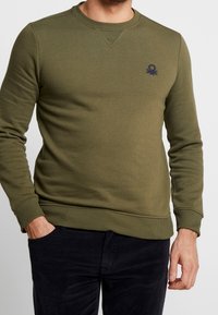 Sudadera verde oliva con cuello redondo, puños y dobladillo acanalados, que presenta un pequeño logo navy en el pecho. Textura suave y diseño ajustado.