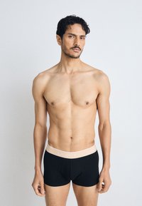 Caleçons noirs à boxer avec une ceinture beige clair portant le logo Lacoste. Tissu lisse, design ajusté, longueur mi-cuisse.