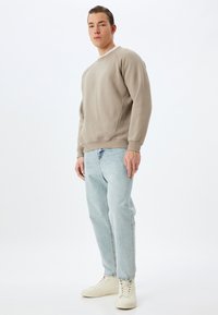 Felpa beige chiaro con polsini e orlo a coste, abbinata a jeans in denim azzurro chiaro. Modello che indossa scarpe bianche alte.