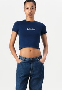 Tengerészkék, rövid ujjú, cropped póló hímzett "Guess Jeans" logóval. Magas derekú farmernadrággal párosítva. Sima anyagú textúra.