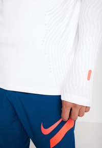 Valkoinen urheilullinen pitkähihainen paita, jossa on tekstuurilinjat ja pieniä rei'ityksiä; yhdistettynä sinisiin housuihin, joissa on suuri oranssi Nike-logo.