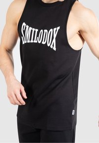 Débardeur noir sans manches en coton, avec un texte blanc "SMILODOX" bien visible sur la poitrine. Texture lisse, coupe régulière.