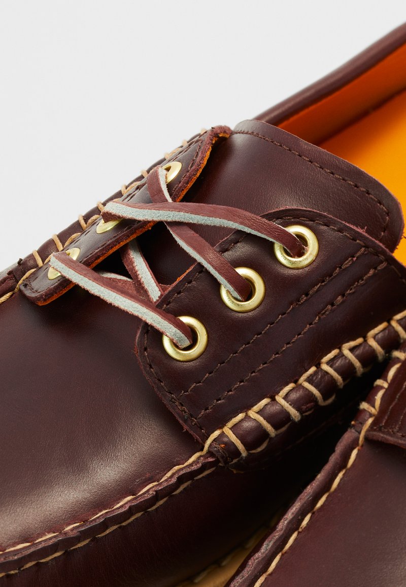 Scarpe da barca in pelle bordeaux con occhielli dorati, cuciture beige e lacci grigio chiaro. L'interno presenta una foderatura arancione brillante.