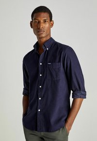 Camisa de botones azul marino de tela de algodón, con cuello, mangas largas remangadas en los puños y un bolsillo en el pecho con etiqueta de logo.