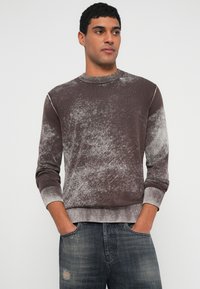 Καφέ και γκρι πουλόβερ με μοτίβο, στρογγυλή λαιμόκοψη, ριμπ μανσέτες και τελείωμα. Φορεμένο με σκούρο τζιν, που αναδεικνύει μια υφή με ανάγλυφη επιφάνεια.