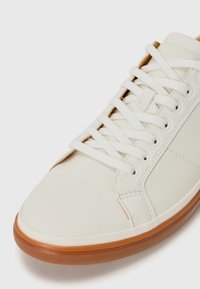 White leather sneaker with a textured finish, featuring a rubber sole and flat white laces. Design includes stitched accents on the side.