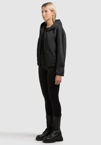 Femme blonde debout, portant une veste à capuche noire, des leggings noirs et des bottes de combat noires à lacets, sur un fond clair uni.