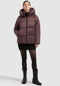 Kvinne iført en brun dunjakke med stor krage, svart miniskjørt, gjennomsiktige svarte strømpebukser og svarte ankelstøvler, stående mot en grå bakgrunn.