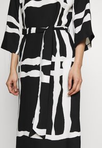 Abito a motivi bianco e nero con maniche ampie, vita con cintura e tessuto fluido. Presenta audaci disegni geometrici a pennellate.