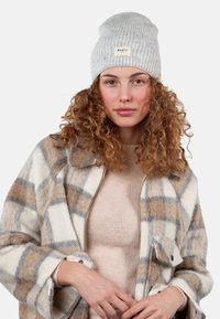 Grey knitted beanie with a label, combined with a beige sweater and a plaid fleece jacket in cream, brown, and blue tones.