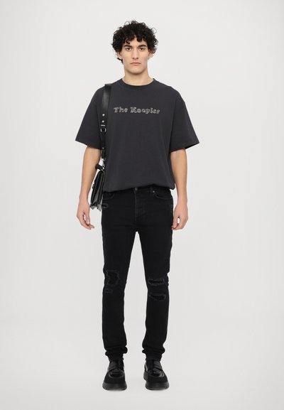 Camiseta gráfica negra con el texto "El Guardián", combinada con jeans negros desgastados y zapatos negros. El modelo sostiene un bolso de hombro negro.