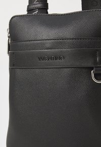 Sac à main noir texturé en matériau synthétique, doté d'un logo bien en vue, d'une bretelle lisse et d'un détail en métal argenté.