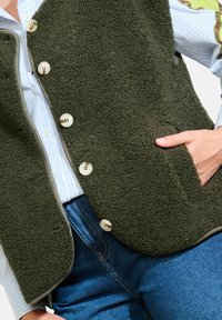 Groen, gestructureerd fleece vest met vijf crèmekleurige knopen en een zijzak, gedragen over een lichtblauwe gestreepte blouse en donkere spijkerbroek.