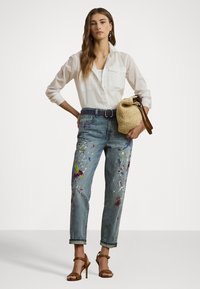 Lightweight white shirt, blue denim jeans with colorful paint splatters, and brown strappy heels; accessorized with a woven bag.