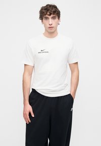 Baltas medvilninis marškinėlis su apvalia iškirpte, trumpomis rankovėmis ir juodu užrašu „NIKE OFFICIAL“ ant krūtinės, derinamas su juodomis sportinėmis kelnėmis.