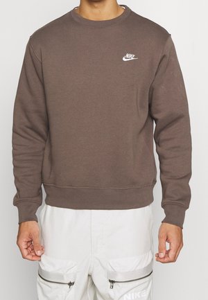 Osoba nosząca brązową bluzę Nike z białym logo na piersi oraz białe spodnie z kieszeniami na suwak i oznakowaniem Nike na udzie.